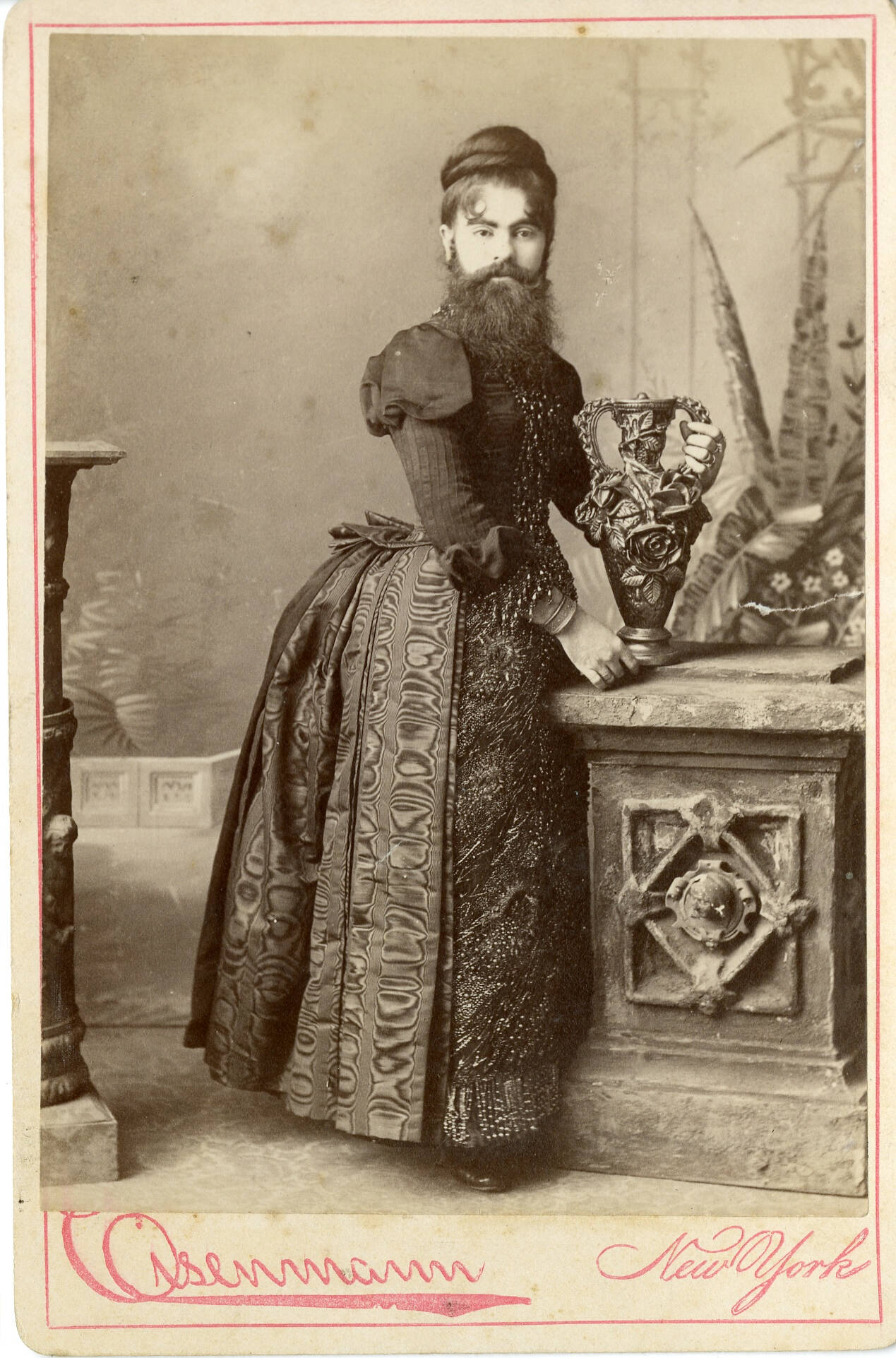Black and white historic cabinet card photograph of a bearded woman in a long dark dress touching a vase on a column.